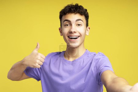 Téléchargez les photos : Portrait de beau garçon souriant, 15 ans influenceur enregistrement vidéo, ayant appel vidéo, portant élégant t-shirt décontracté violet regardant la caméra isolée sur fond jaune. Concept de blogging - en image libre de droit