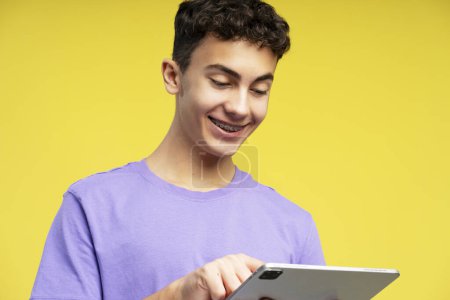Téléchargez les photos : Portrait d'un garçon attrayant et souriant, adolescent portant un t-shirt violet, tenant une tablette numérique isolée sur fond jaune, gros plan. Concept de technologie, éducation en ligne - en image libre de droit