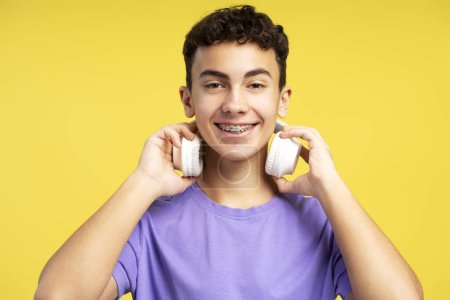 Photo for Portrait of handsome smiling boy with braces wearing headphones listening to music, looking at camera isolated on yellow background. Technology concept - Royalty Free Image