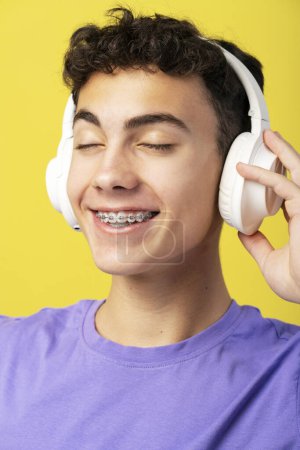 Téléchargez les photos : Portrait rapproché d'un garçon souriant avec des accolades écoutant de la musique dans des écouteurs jouissant avec les yeux fermés isolés sur fond jaune. Concept de publicité - en image libre de droit