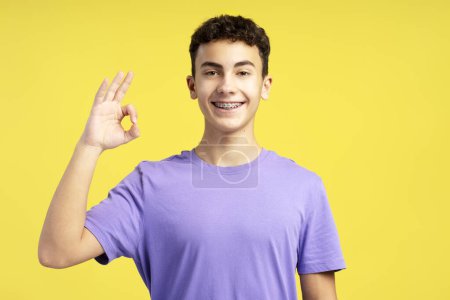 Foto de Retrato de niño guapo sonriente con frenos dentales que usan una camiseta púrpura casual, mostrando un signo aceptable con los dedos mirando a la cámara aislada sobre un fondo amarillo. Concepto de publicidad - Imagen libre de derechos