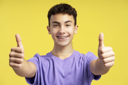Foto de Retrato de niño guapo sonriente, adolescente de 15 años con tirantes dentales con elegante camiseta púrpura informal que muestra los pulgares hacia arriba mirando a la cámara aislada en el fondo amarillo. Concepto de publicidad - Imagen libre de derechos