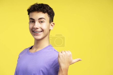 Foto de Niño guapo y sonriente, adolescente con frenos dentales apuntando con el dedo al espacio de copia, mirando a la cámara de pie aislada sobre fondo amarillo. Concepto ortodoncista - Imagen libre de derechos