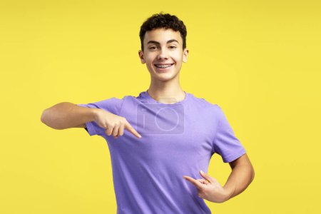 Photo for Portrait of smiling attractive teenager wearing dental braces casual purple t shirt pointing finger at himself, looking at camera isolated on yellow background. Advertisement concept - Royalty Free Image