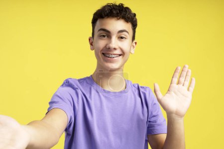 Téléchargez les photos : Portrait d'un adolescent excité souriant avec des appareils dentaires agitant la main, portant un t-shirt violet décontracté ayant un appel vidéo regardant la caméra debout isolée sur fond jaune. Concept de blogging - en image libre de droit