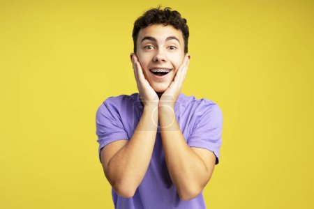 Foto de Retrato de un atractivo adolescente excitado con tirantes mirando a la cámara, usando una elegante camiseta púrpura de pie aislada sobre un fondo amarillo. Concepto de compras - Imagen libre de derechos