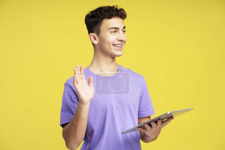 Téléchargez les photos : Portrait d'un adolescent élégant et souriant avec des orthèses dentaires portant des vêtements décontractés tenant une tablette numérique agitant la main en regardant loin isolé sur fond jaune. Concept technologique - en image libre de droit