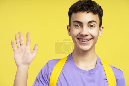 Téléchargez les photos : Portrait de garçon souriant, adolescent, étudiant avec des orthèses dentaires portant une chemise violette élégante et un sac à dos agitant la main en regardant la caméra isolée sur fond jaune. Concept d'éducation - en image libre de droit