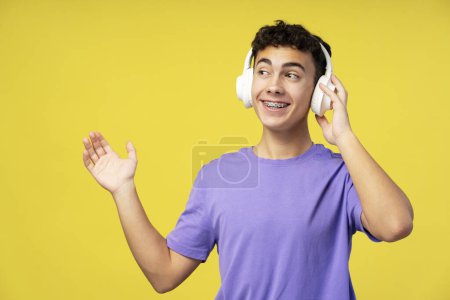 Téléchargez les photos : Portrait d'un adolescent souriant portant un t-shirt violet décontracté regardant loin en écoutant de la musique dans des écouteurs montrant la main isolée sur fond jaune. Concept technologique - en image libre de droit