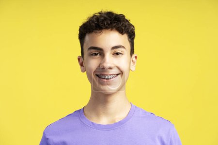 Téléchargez les photos : Portrait rapproché d'un garçon confiant et souriant avec des orthèses dentaires sur les dents regardant la caméra isolée sur fond jaune. Soins de santé, concept orthodontique - en image libre de droit