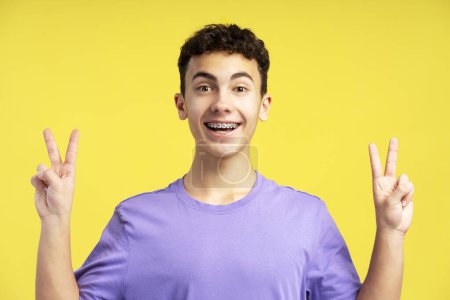 Téléchargez les photos : Adolescent souriant avec des appareils dentaires montrant signe de victoire en regardant la caméra isolée sur fond jaune. Émotions positives, soins de santé, concept orthodontique - en image libre de droit