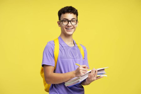 Téléchargez les photos : Intelligent garçon d'école souriant portant des lunettes tenant des livres prenant des notes isolées sur fond jaune. Étudiant avec sac à dos étudiant, apprendre la langue en regardant la caméra. Concept d'éducation - en image libre de droit