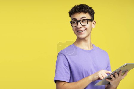 Foto de Retrato inteligente niño sonriente de la escuela con anteojos usando tableta digital, estudio, comunicación en línea mirando hacia otro lado aislado en el fondo amarillo, espacio de copia. Concepto de educación en línea - Imagen libre de derechos