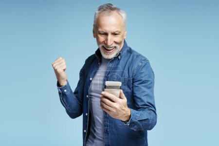 Téléchargez les photos : Portrait d'un homme mûr aux cheveux gris souriant tenant un téléphone portable, regardant une vidéo faisant un geste de victoire isolé sur fond bleu. Homme senior vérifiant le courrier. Concept technologique - en image libre de droit