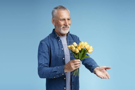 Téléchargez les photos : Portrait d'un séduisant homme âgé souriant portant des vêtements décontractés élégants tenant un bouquet de fleurs donnant des tulipes jaunes de printemps regardant la caméra. Concept de Mars 8, datant - en image libre de droit