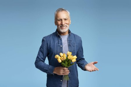 Téléchargez les photos : Portrait d'un séduisant homme âgé souriant portant des vêtements décontractés élégants tenant un bouquet de fleurs, des tulipes jaunes de printemps regardant l'appareil photo. Concept de journée de la femme - en image libre de droit