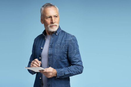 Photo for Portrait of serious gray haired senior man, businessman wearing stylish casual clothes looking away, holding notebook, taking notes isolated on blue background. Business concept - Royalty Free Image
