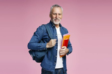 Photo for Portrait of smiling senior man, mature student holding backpack and books looking at camera isolated on pink background. Education concept - Royalty Free Image