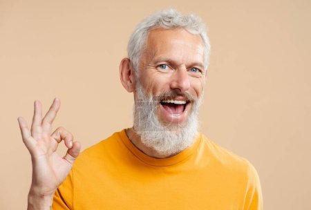 Téléchargez les photos : Beau sourire homme de 50 ans avec des cheveux gris élégant portant t-shirt orange décontracté montrant ok, regardant la caméra isolée en arrière-plan. Soins de santé, concept de traitement dentaire - en image libre de droit