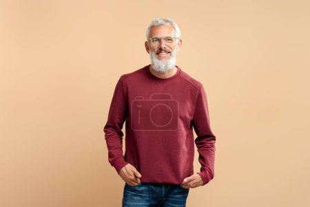 Photo for Handsome smiling mature man, bearded gray haired hipster wearing stylish eyeglasses holding hands in pockets looking at camera isolated on beige background. Successful business - Royalty Free Image