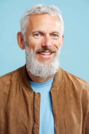 Téléchargez les photos : Portrait bel homme mature aux cheveux gris souriant, hipster barbu portant une veste en cuir élégante regardant la caméra isolée sur fond bleu - en image libre de droit