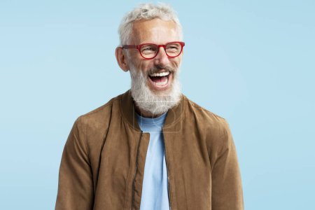 Foto de Auténtico retrato guapo feliz de mediana edad hombre de pelo gris con elegantes gafas hipster rojas mirando hacia otro lado y riendo aislado sobre fondo azul. Emociones, concepto de estilo de vida positivo - Imagen libre de derechos