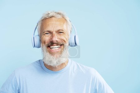 Foto de Hombre maduro guapo feliz, hipster barbudo de pelo gris escuchando música en auriculares inalámbricos aislados sobre fondo azul. Estilo de vida positivo, concepto de tecnología - Imagen libre de derechos
