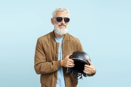 Foto de Hombre maduro guapo serio fresco, motorista barbudo de pelo gris con gafas de sol elegantes, chaqueta de cuero marrón que sostiene el casco de la motocicleta aislado sobre fondo azul - Imagen libre de derechos