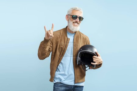 Photo for Handsome smiling mature man, cool gray haired biker wearing stylish sunglasses, brown leather jacket holding motorcycle helmet isolated on blue background. Positive lifestyle, freedom concept - Royalty Free Image