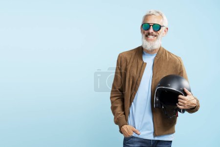 Foto de Guapo sonriente hombre maduro fresco, motero barbudo de pelo gris sosteniendo casco de motocicleta aislado sobre fondo azul - Imagen libre de derechos