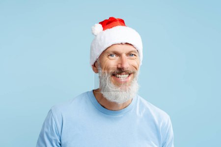 Photo for Closeup portrait handsome smiling mature man wearing red Santa hat looking at camera celebration Christmas isolated on blue background. Holiday concept - Royalty Free Image