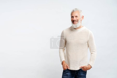 Téléchargez les photos : Beau barbu homme aux cheveux gris portant chandail élégant tenant la main dans des poches isolées sur fond blanc - en image libre de droit