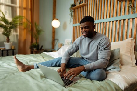 Téléchargez les photos : Souriant étudiant afro-américain attrayant en utilisant un ordinateur portable, se préparant à l'examen assis sur un lit confortable dans une chambre confortable à la maison. Concept d'emploi à distance, éducation en ligne - en image libre de droit