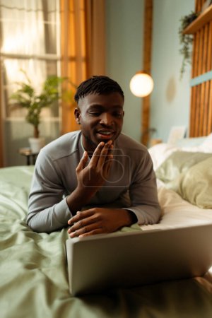 Foto de Retrato de un atractivo hombre africano que se comunica con el lenguaje de señas, tiene videoconferencia, usa computadora portátil, hace gestos mientras está acostado en la cama, en casa. Concepto de comunicación - Imagen libre de derechos