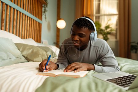 Téléchargez les photos : Portrait d'un étudiant afro-américain souriant portant un casque écoutant une conférence, tenant un stylo, prenant des notes, utilisant un ordinateur portable. ? acceptation de l'éducation en ligne, préparation à l'examen - en image libre de droit