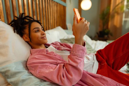 Téléchargez les photos : Portrait d'heureuse femme afro-américaine souriante portant des vêtements colorés élégants tenant téléphone portable prenant selfie tout allongé sur le lit à la maison. Concept de technologie en ligne, achats - en image libre de droit