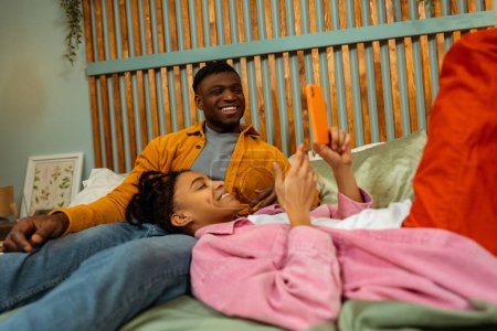 Téléchargez les photos : Portrait d'un homme et d'une femme afro-américains souriants, beau jeune couple, couché sur le lit ensemble, utilisant le téléphone portable, regardant la vidéo. Concept de relations, amour - en image libre de droit