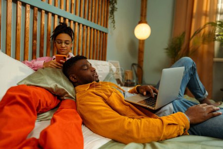 Téléchargez les photos : Portrait de jeune homme et femme afro-américain moderne, pigistes portant des vêtements décontractés colorés élégants couchés sur le lit, à l'aide d'un ordinateur portable et d'un téléphone mobile, à la maison. Concept de technologie en ligne - en image libre de droit