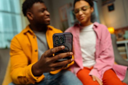 Photo for Portrait of handsome African American man and woman holding mobile phone, watching video together, selective focus on hand, at home. Online technology concept - Royalty Free Image