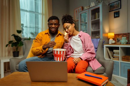 Téléchargez les photos : Jeune couple souriant, homme et femme afro-américain portant des vêtements décontractés colorés élégants assis sur un canapé confortable, mangeant du pop-corn, utilisant un ordinateur portable, regardant une vidéo, un film. Concept de détente - en image libre de droit