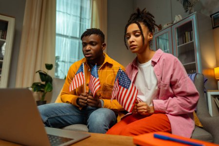 Téléchargez les photos : Portrait d'un couple afro-américain sérieux et coûteux tenant des drapeaux américains, utilisant un ordinateur portable, attendant les résultats de l'élection présidentielle, regardant une vidéo alors qu'il était assis sur le canapé à la maison. Concept du jour d'élection - en image libre de droit