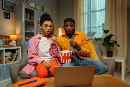 Photo for Excited, shocked young couple, African American man and woman wearing stylish colorful casual clothes sitting on comfortable sofa, eating popcorn, using laptop, watching movie. Relaxation concept - Royalty Free Image
