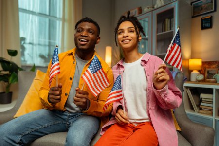Téléchargez les photos : Portrait d'un beau couple afro-américain souriant, de jeunes supporters américains tenant des drapeaux américains se réjouissant des résultats des élections assis chez eux. Concept d'élection présidentielle américaine, célébration - en image libre de droit