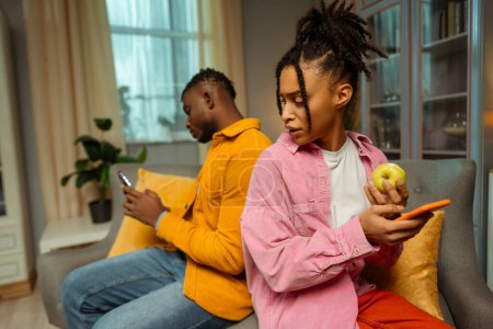 Téléchargez les photos : Portrait d'une femme afro-américaine attrayante qui porte des vêtements colorés tenant un téléphone portable et une pomme, assise à côté d'un jeune homme sur un canapé à la maison. Concept de relation, technologie en ligne - en image libre de droit