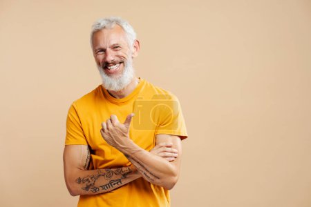 Téléchargez les photos : Portrait d'homme mûr souriant attrayant, hipster barbu aux cheveux gris portant un t-shirt jaune, tenant les bras croisés pointant du doigt sur l'espace de copie isolé sur fond blanc. Concept de publicité - en image libre de droit