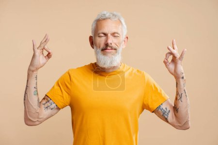 Photo for Portrait of handsome gray haired middle aged man wearing stylish yellow t shirt with eyes closed meditating, standing isolated on beige background. Concept of balance, harmony - Royalty Free Image