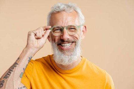 Foto de Sonriente hombre maduro exitoso, con gafas con estilo y camiseta amarilla casual, mirando a la cámara, de pie aislado sobre fondo beige. Concepto de visión - Imagen libre de derechos
