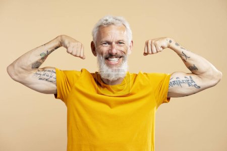 Photo for Portrait of smiling mature man wearing casual yellow t shirt showing biceps, tattooed hand, looking at camera, standing isolated on beige background. Sport concept - Royalty Free Image