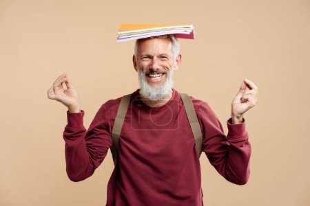 Téléchargez les photos : Portrait d'un homme barbu souriant, étudiant portant un sac à dos, tenant des cahiers et des livres sur la tête, s'amusant, regardant la caméra, gesticulant debout isolé sur fond beige. Concept d'éducation - en image libre de droit