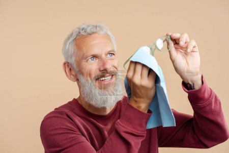 Téléchargez les photos : Portrait d'un homme mûr coûteux portant un pull portant des lunettes, essuyant avec une serviette, debout isolé sur fond beige. Concept de soins de la vue - en image libre de droit